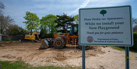 heavy machinery works in the background with a sign that says 'please pardon our appearance' as new playground equipment will be installed