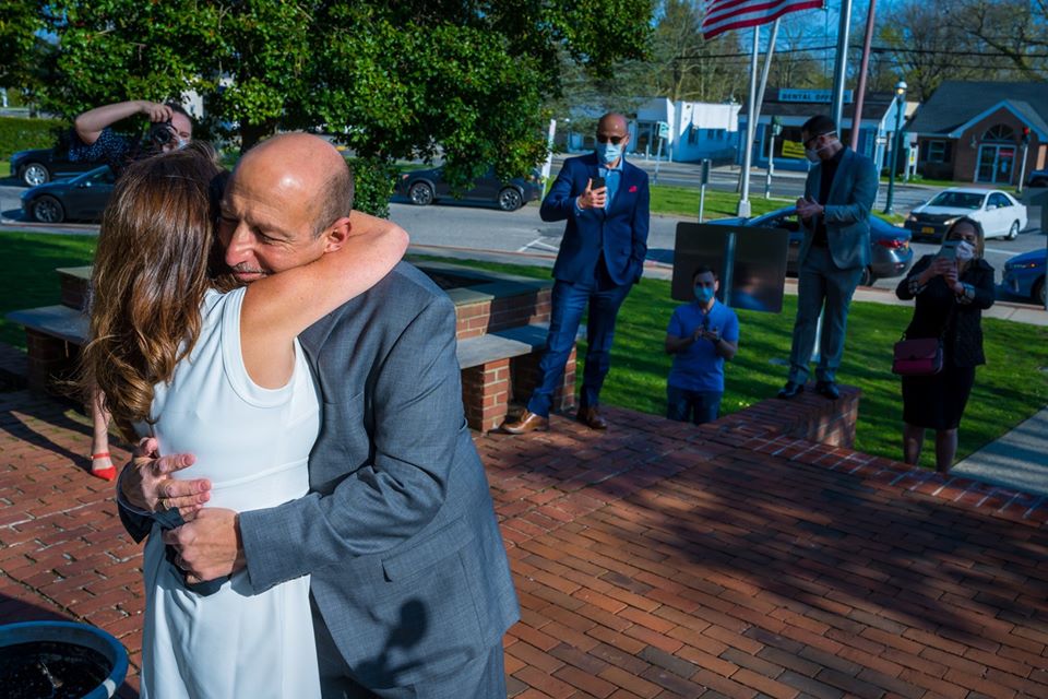 Bride and Groom hug while witnesses can be seen behind them 6 feet apart