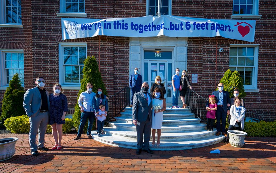 Bride and Groom and Wedding party stand in familial groups, 6 feet apart all in front of the Town Hall steps and the banner "we're in this together...but 6 feet apart" 