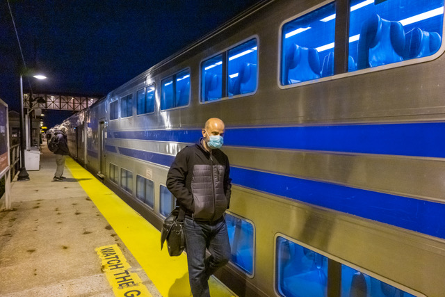 A man in a mask walks beside the double decker train, hands in pocket, during early hours. No one near him.