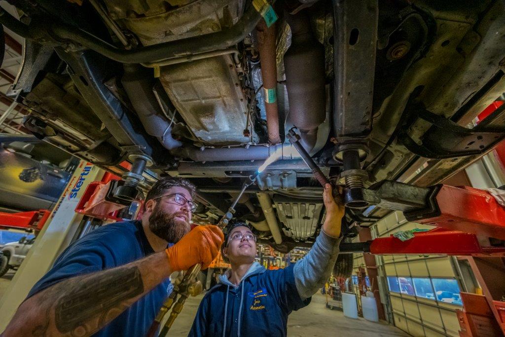 A student is shown the process of welding under the frame of a raised car by an experienced technician.