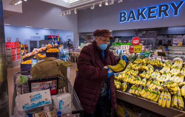 A woman in a mask and gloves picks bananas from the stocked produce section in a supermarket. No one near her.