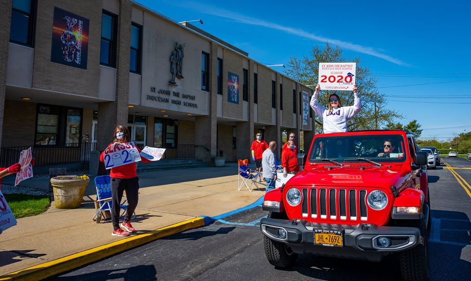 Fire engine red jeep with student holding sign up through moon roof drives past teachers in ppe