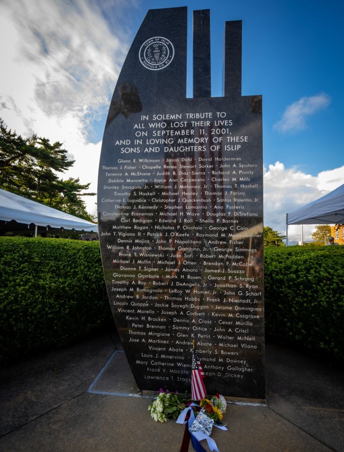 A front on photo of the Islip September 11th Memorial, the American flag, draped over main street reflecting in the dark polished stone as the names of the fallen show etched in white