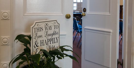 An image of the Town Board room doors cracked slightly open with a sign in the foreground saying, this way to love, laughter, and happily ever after.