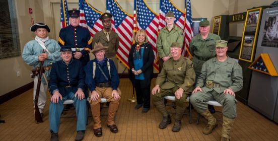 Supervisor Carpenter stands infront of a row of American Flags with military re-enactors beside her and sitting in front.