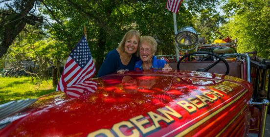 Supervisor Carpenter standing at a vintage Ocean Beach Fire engine next to Parade honoree, Natalie Katz Rogers, sun lit trees and american flags blowing beside them.