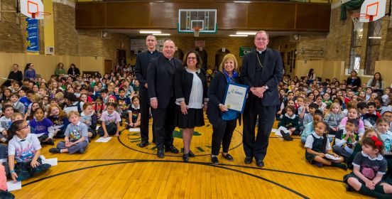  The Bishop, Supervisor Carpenter and School Officials pose for a photo in the middle of a gymnasium as scores of school look on, seated beside them.