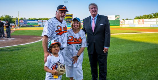 Supervisor Carpenter stands with her grandson, Ducks Coach, as well as Ducks Owner Frank Boulton on the ballfield in the shadow of the setting sun, having just thrown out the first pitch.