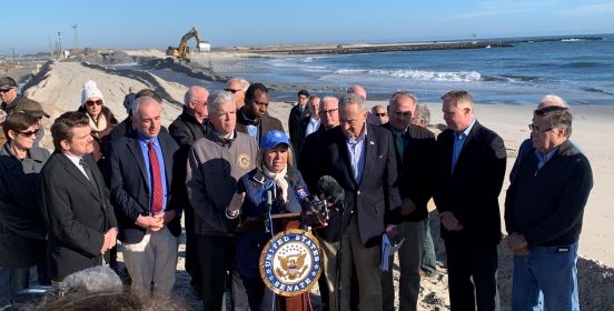 supervisor Carpenter speaks at the podium, addressing the media as County Executive Bellone and Senator Schumer and local officials stand beside her, the shoreline and construction vehicles seen in the distance behind her.