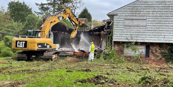 Construction vehicle begins demolition on property as workers spray debris with hose.