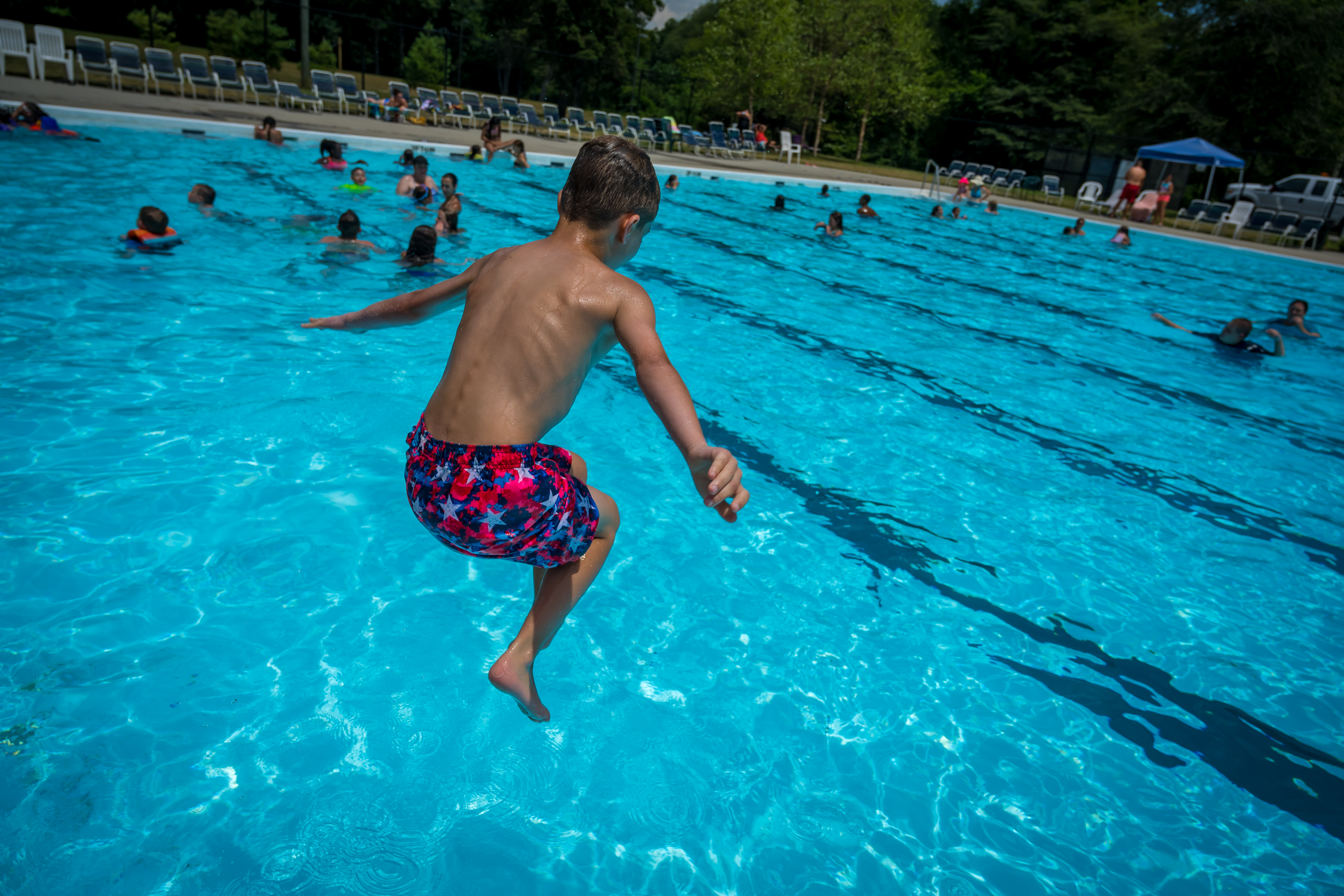A young boy seen with arms outstretched and knees tucker as he hovers over the pool in mid air, about to crash into the blue sparkling water