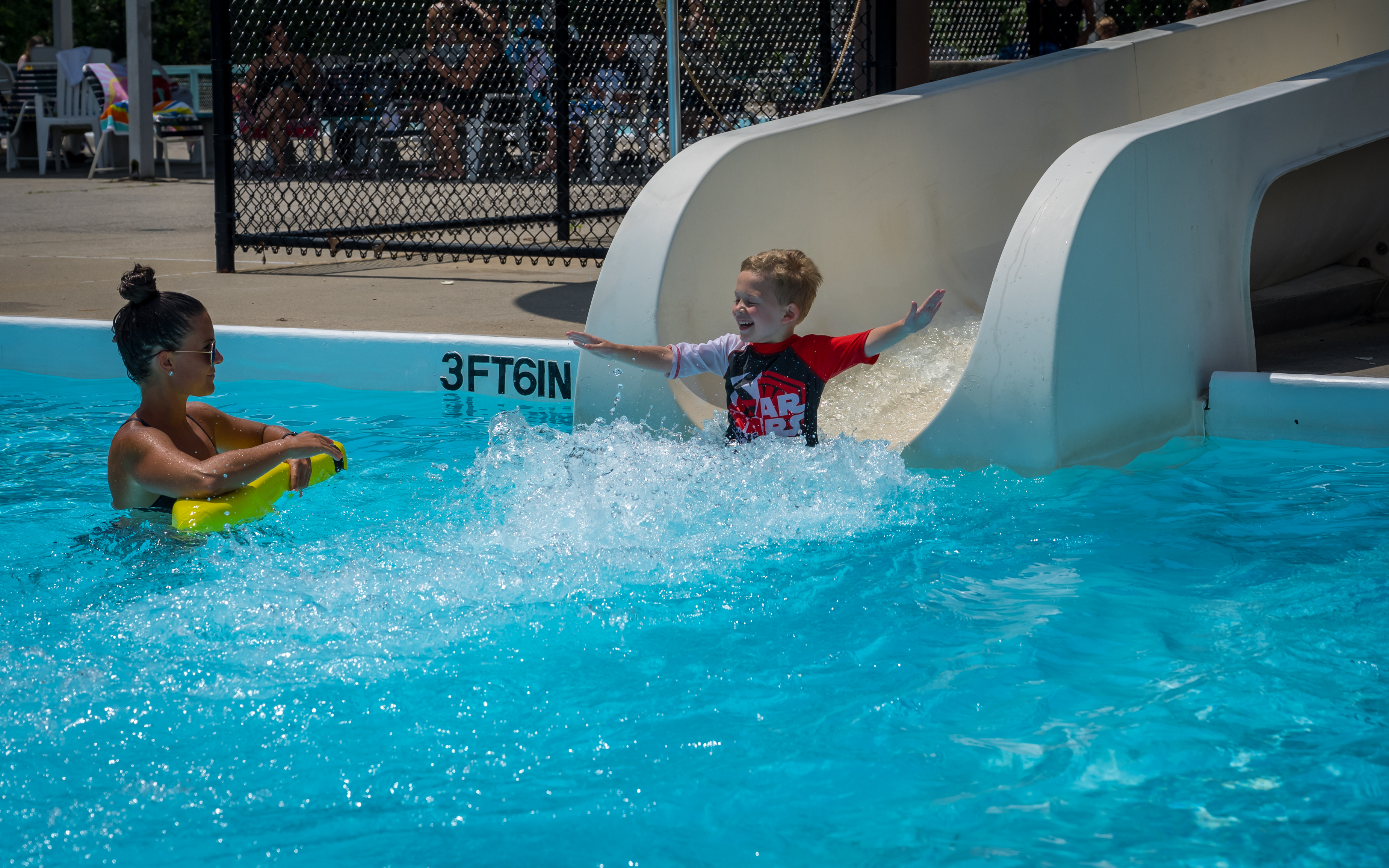 Young boy with a big smile on his face and arms out like an airplane as he cruises toward his descent into the cool blue pool, a lifeguard to his left ready to catch him if needed.